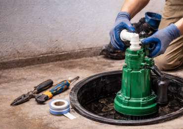 Technician installing or repairing a green sump pump inside a basement sump pit with plumbing tools nearby.